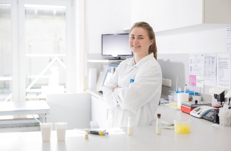 Eine Mitarbeiterin im Labor trägt einen weißen Kittel, steht an einem Tisch mit Proben und arbeitet an der Qualitätskontrolle von Milchprodukten.